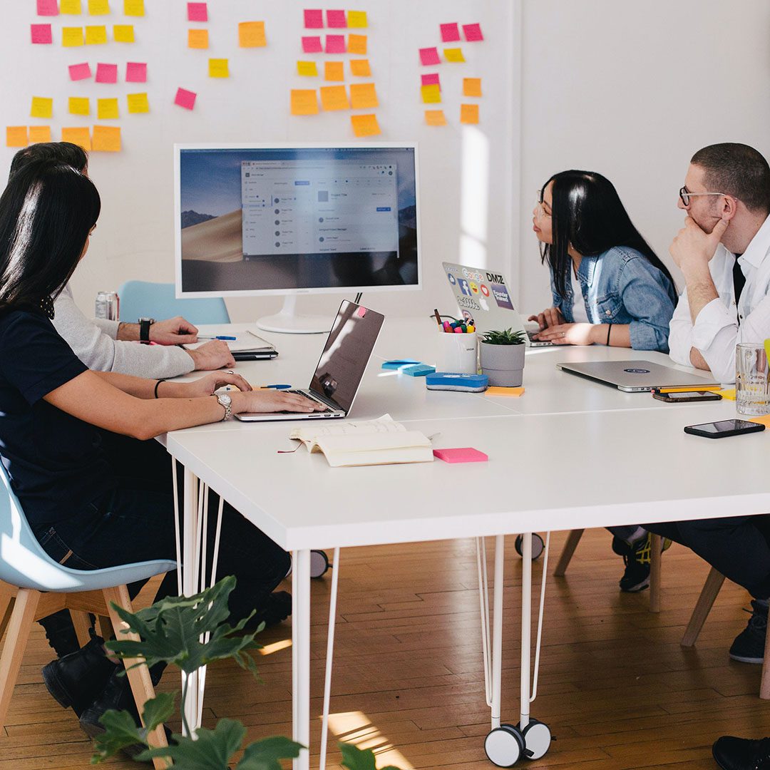 Four people sat around a desk in a meeting room environment all looking at a screen at the end of the table with laptops upon the desk and colourful post it notes on the wall behind discussing their company's need for SaaS solutions and support from Cloud Assist Group.