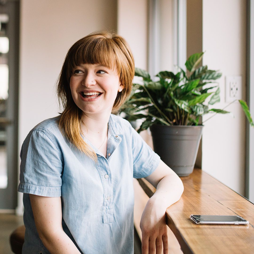 A woman sat at a breakfast bar style desk at a window is looking behind her smiling with her phone in front of her on the desk knowing her company has implemented services from Cloud Assist Group to help their growth through SaaS solutions and support.