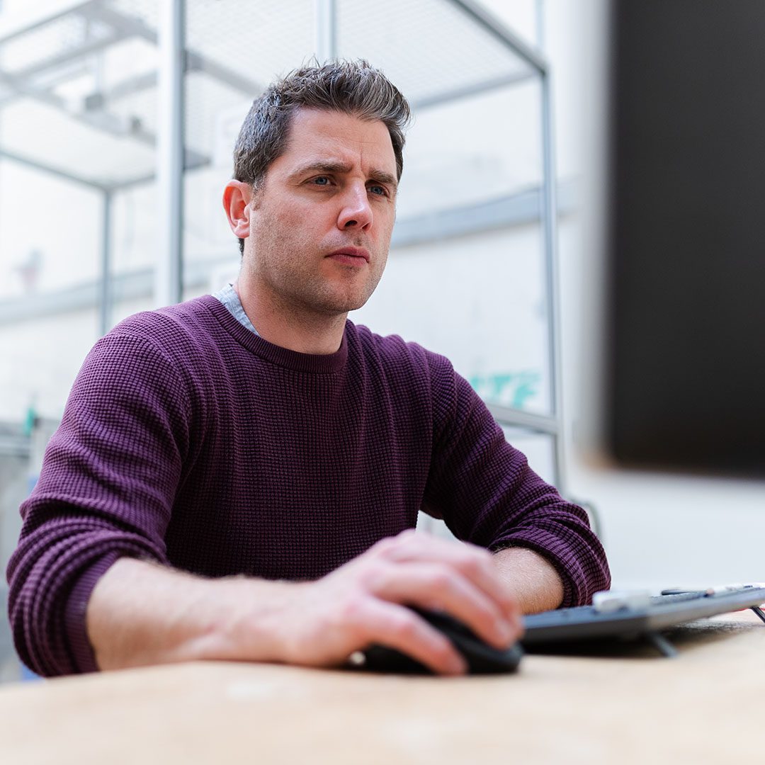 Man sat at a desk in office environment concentrating on what is on his screen looking for SaaS solutions and support from Cloud Assist Group, whilst his right hand operates a computer mouse.
